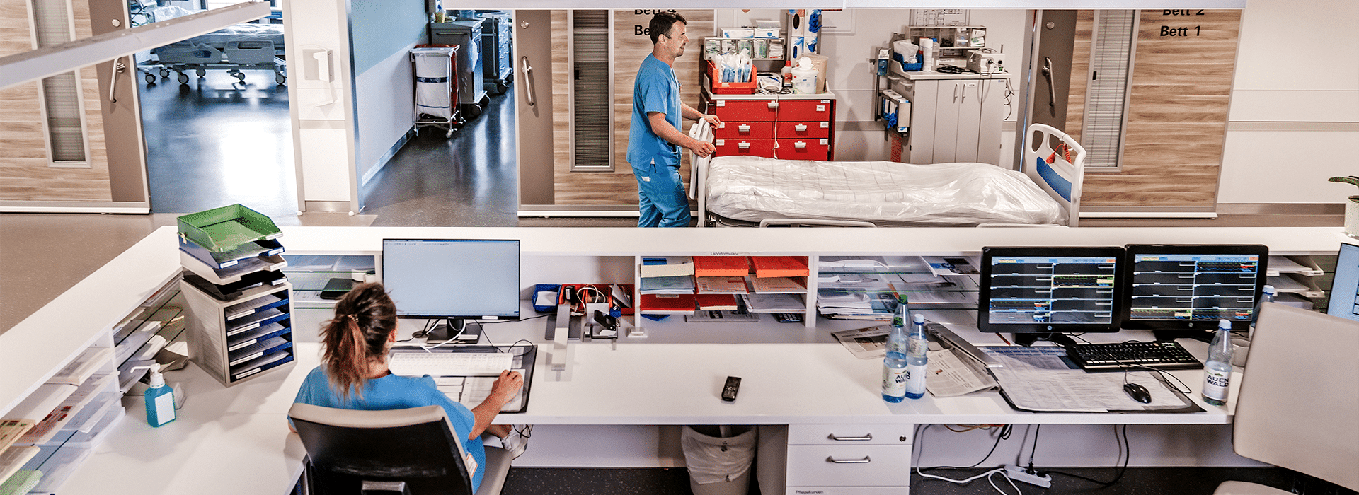 Blick in eine Intensivstation, eine weibliche Pflegefachkraft sitzt an einem Tresen, eine männliche Pflegfachkraft schiebt ein Bett