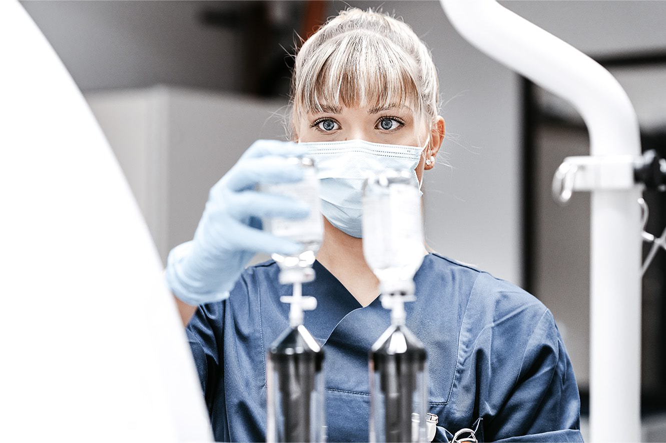 Medizinische weibliche Fachkraft im blauen Kasack mit Mundschutz fokussiert auf eine medizinische Gerätschaft. Sie trägt eine Maske.