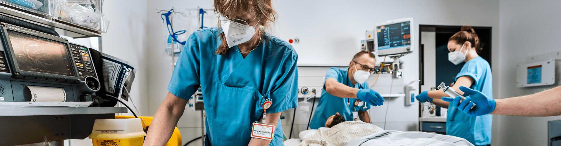 Patientenzimmer mit vielen Monitoren und Bett mit Patient. Drei medizinische Fachkräfte mit Mundschutz im blauen Kasack versorgen den Patienten.