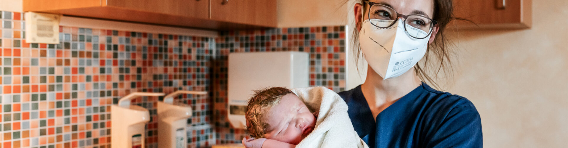 Medizinische, weibliche Fachkraft im blauen Kasack mit Brille und Mundschutz hält ein neugeborenes Baby das in ein weißes Handtuch gewickelt ist. Im Hintergrund eine bunte Kachelwand und Desinfektionsspender.