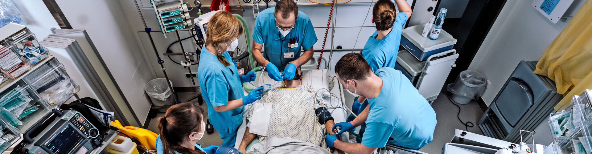 Draufsicht auf ein Patientenbett mit Patient der von fünf medizinischen Fachkräften in blauen Kasacks versorgt wird. Drumherum viele medizinische Geräte und Instrumente.