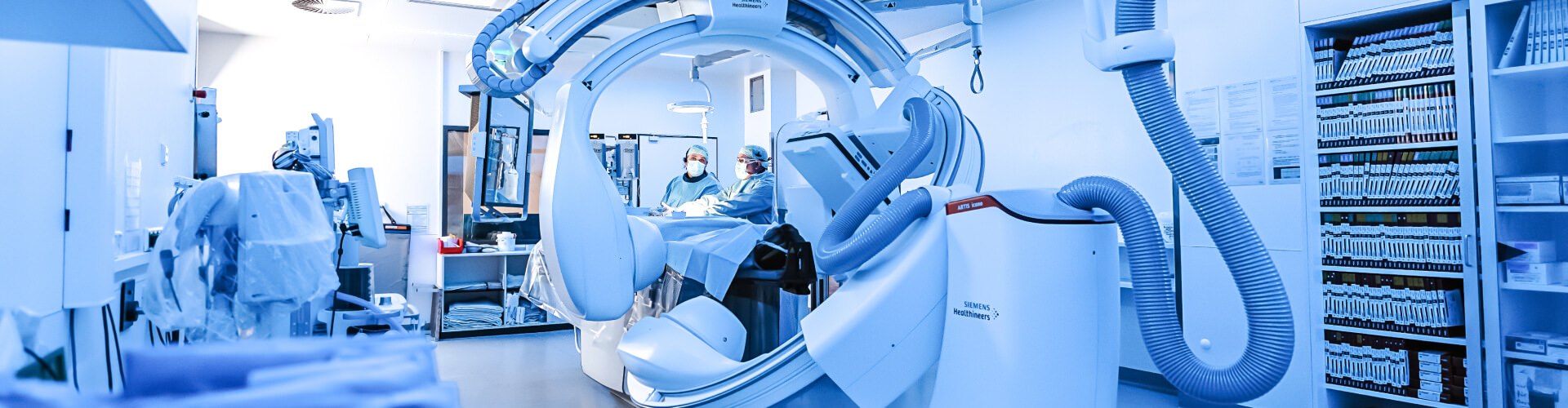Medizinischer Raum in blau Filter getaucht. Vorne ein großes medizinische Gerät zur Bildgebung. Hinten zwei Menschen in OP-Kleidung an einem OP-Tisch. Rechts und links medizinisches Material in Regalen.