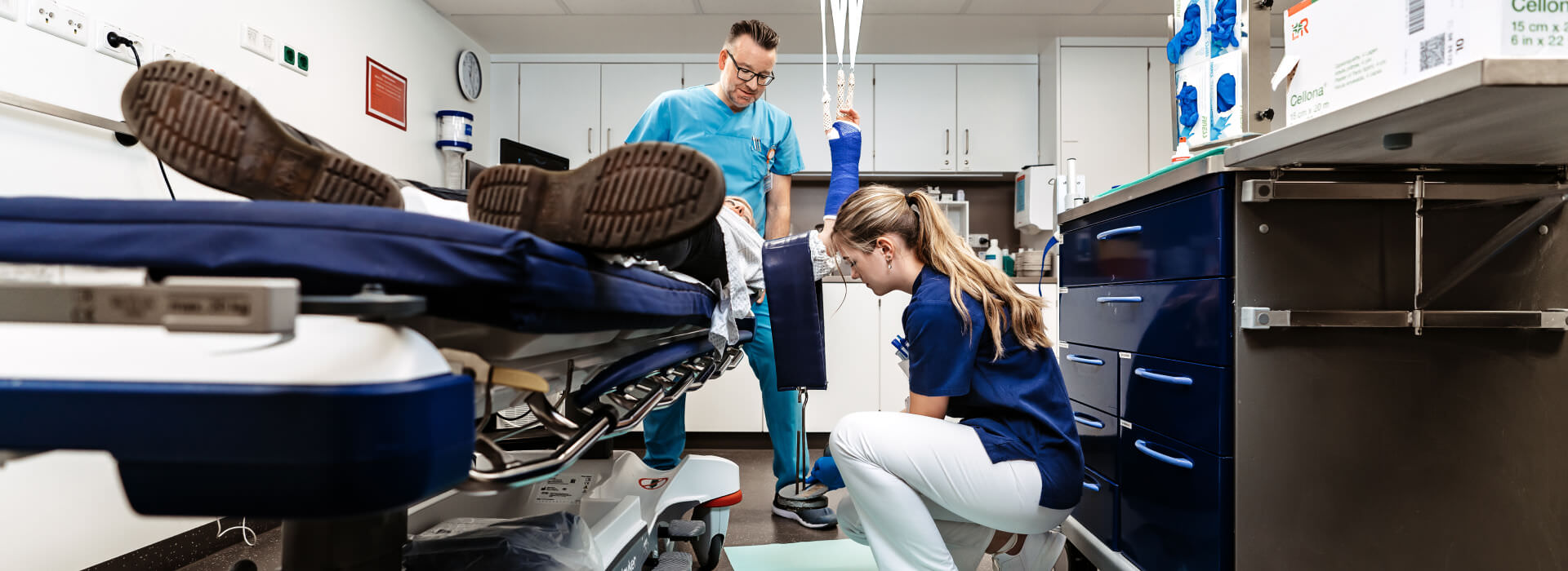 Untersuchungszimmer mit zwei medizinischen Fachkräften im blauen Kasack. Eine kniend vor einer Krankenliege auf der jemand liegt von dem nur die Füße und einen eingegipsten Arm sieht. Rechts ein schwarzer Schrank an der Rückwand ein weißer Schrank.