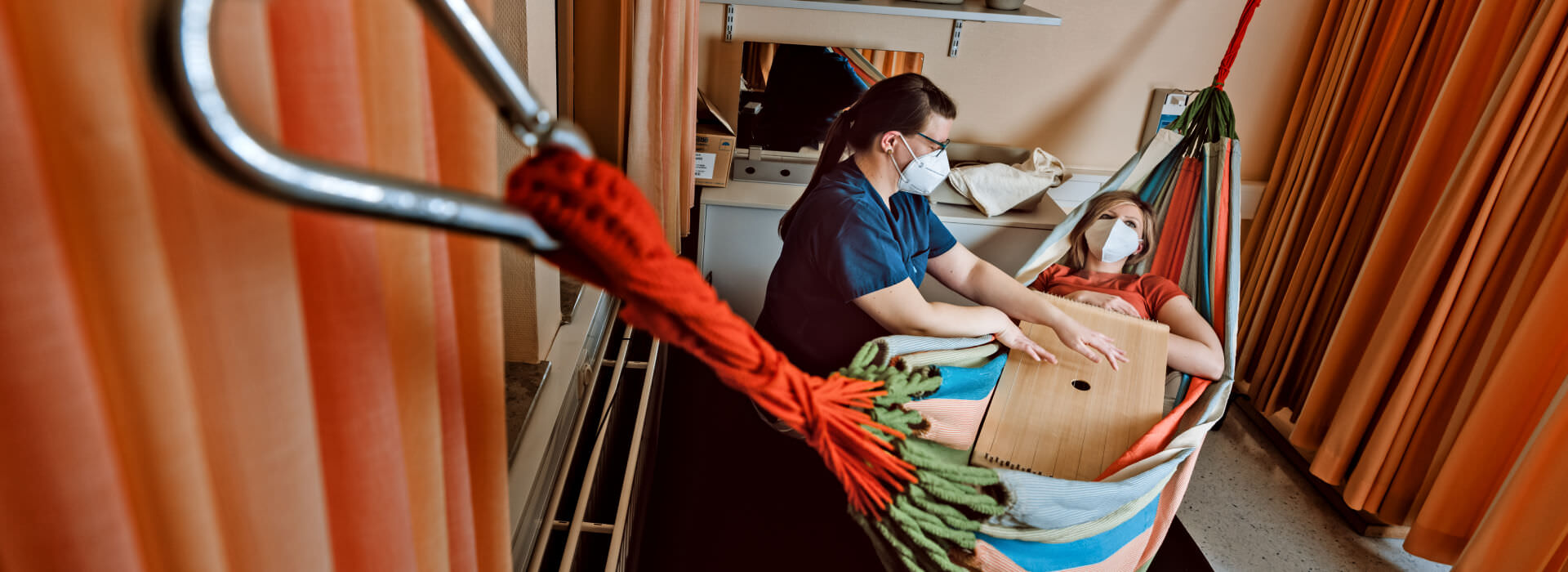Vogelperspektive auf eine weibliche Person in einer Hängematte in Alltagskleidung mit Mundschutz. Daneben eine weibliche medizinische Fachkraft im blauen Kasack mit Mundschutz die ein Musikinstrument auf dem Bauch der liegenden Person spielt. Rechts und links orange-gelbe Vorhänge die den Bereich abschirmen.