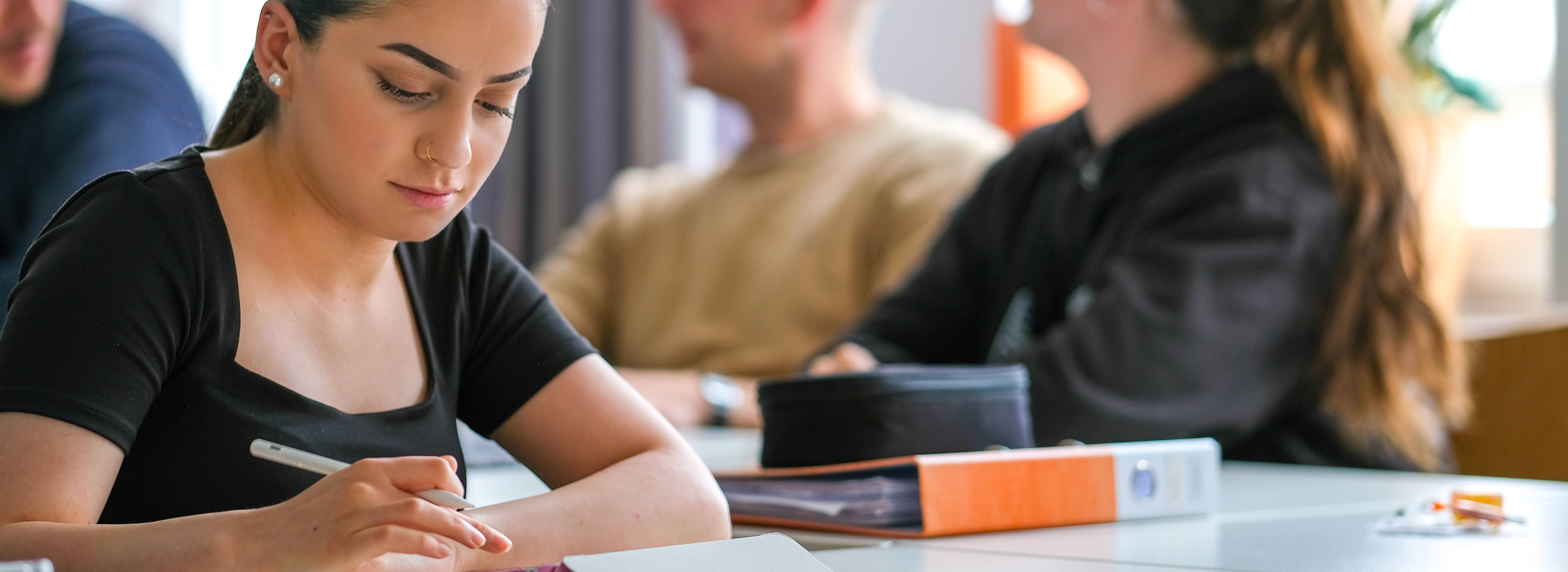 Weibliche Person in Alltagskleidung im Klassenzimmer am Tisch sitzend und auf das vor ihr liegende Tablet blickend. Im Hintergrund verschwommen weitere Personen an Tischen.