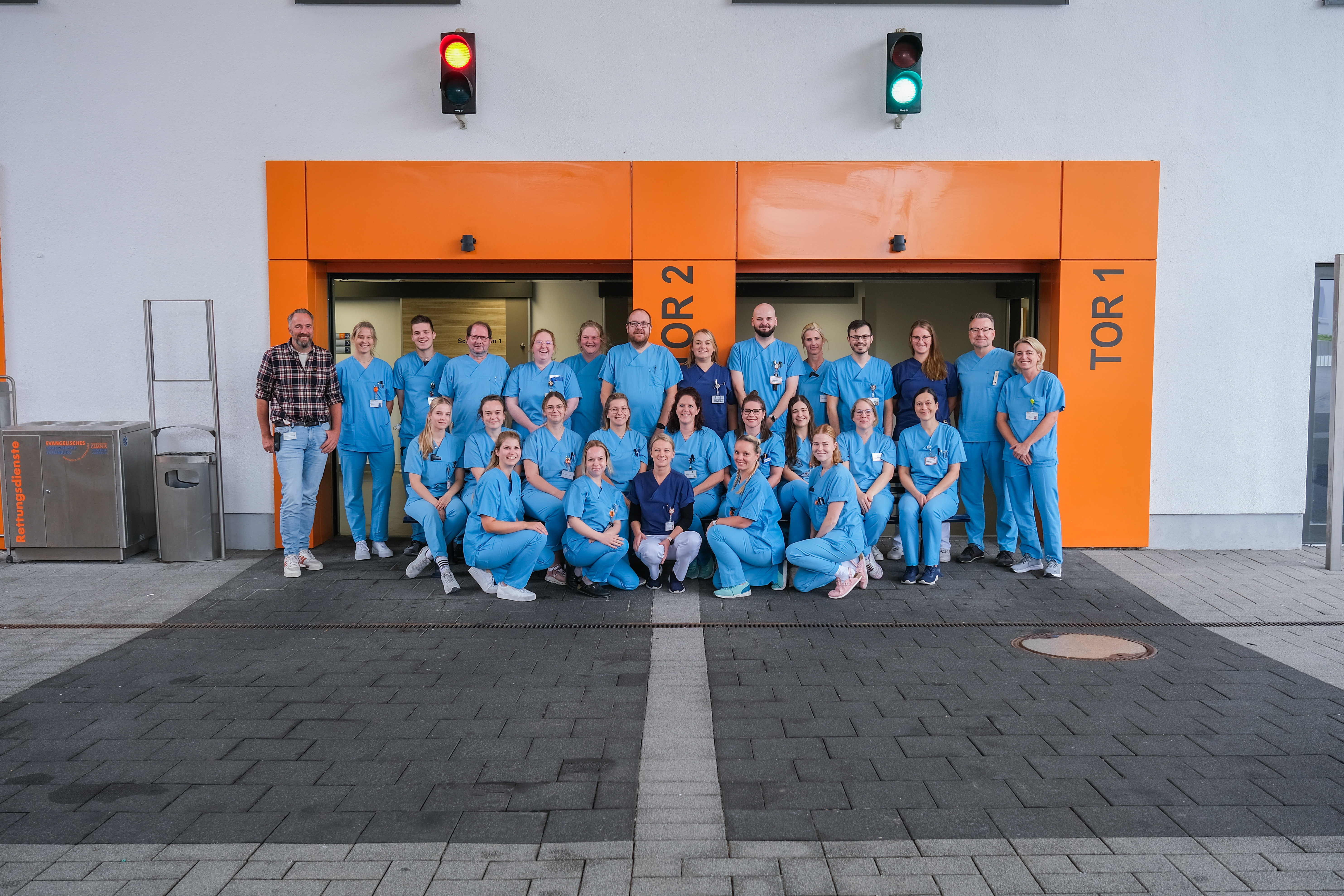 Gruppenfoto mit medizinischem Personal in blauen Kasacks vor den Toren der Notaufnahme. Um die Tore sind große orangene Rahmen mit grauer Beschriftung. Über den Toren Ampeln - eine auf grün eine auf rot.