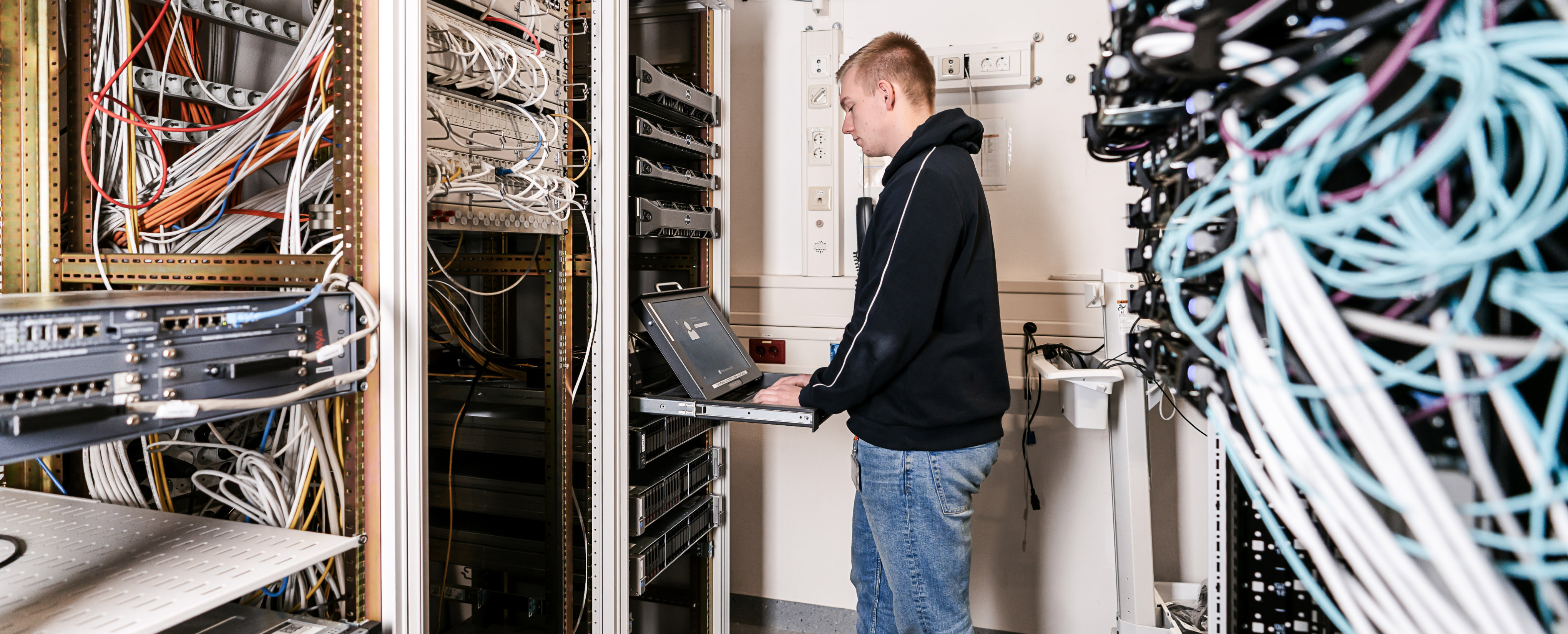 Mann in dunklem Pullover und Jeans in einem Server-Raum vor einem Laptop. Überall sind Kabel.