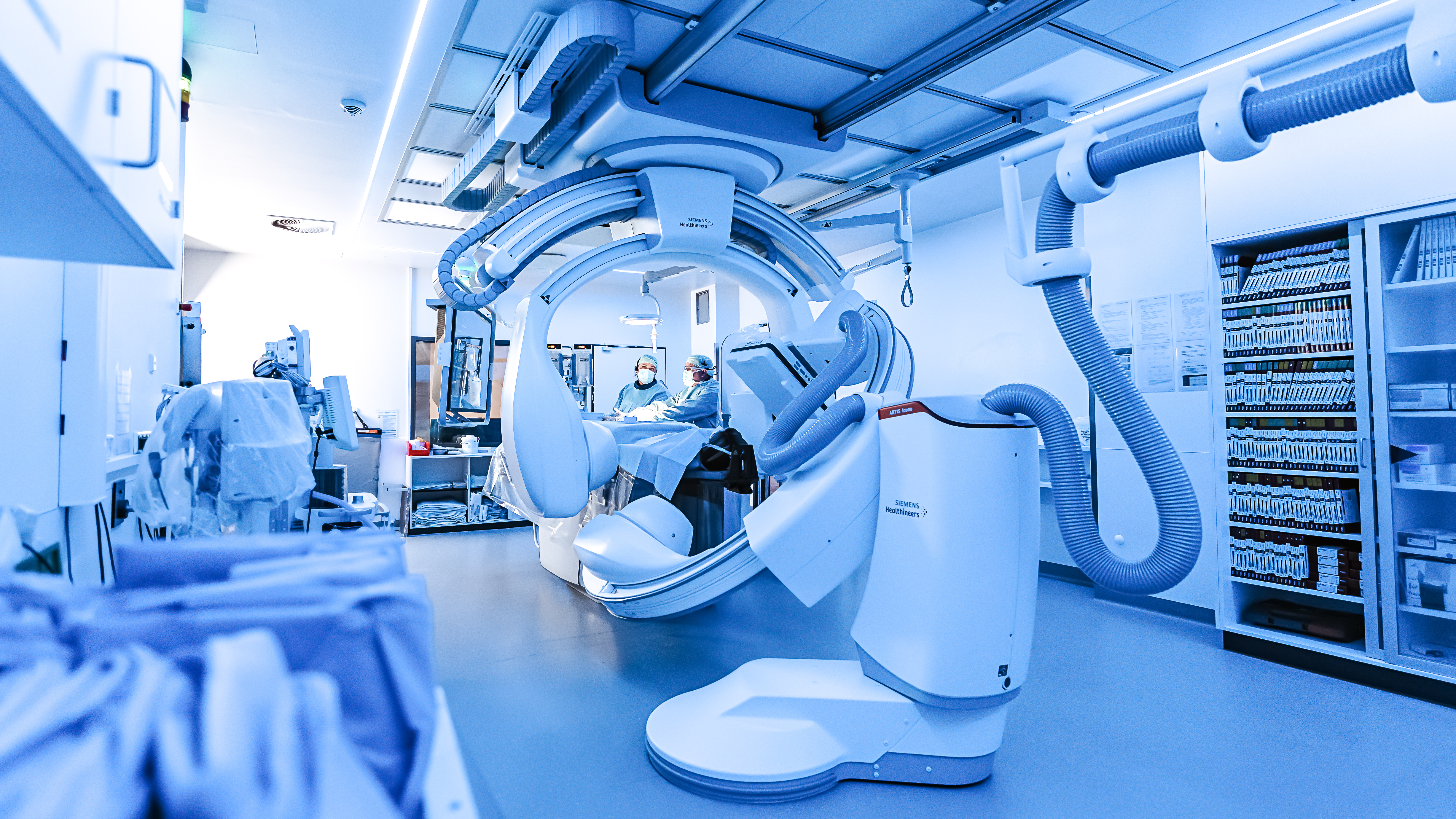 Medizinischer Raum in blau Filter getaucht. Vorne ein großes medizinische Gerät zur Bildgebung. Hinten zwei Menschen in OP-Kleidung an einem OP-Tisch. Rechts und links medizinisches Material in Regalen.