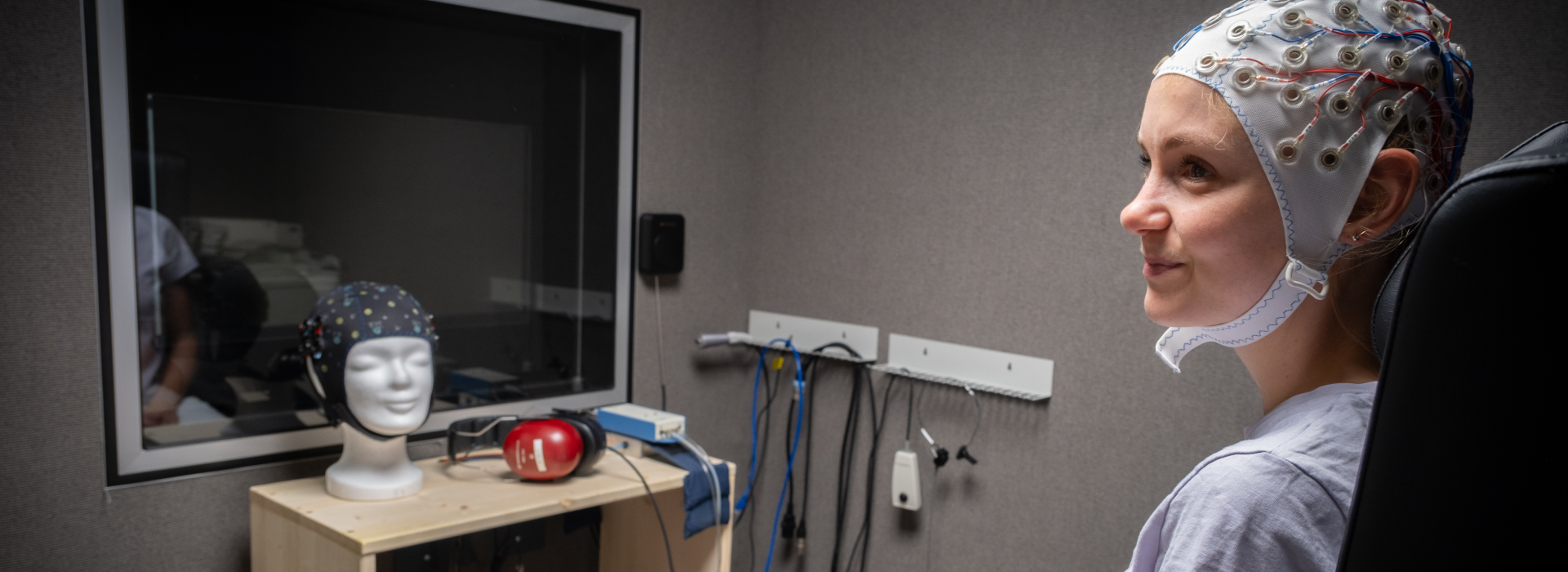 Rechts im Bild eine sitzende Frau mit einer Haube mit Elektroden auf dem Kopf. Links im Bild ein Fenster zum nächsten Raum. Davor eine Regal mit Kopfhörern und einem Modellkopf mit einer Haube mit Elektroden.