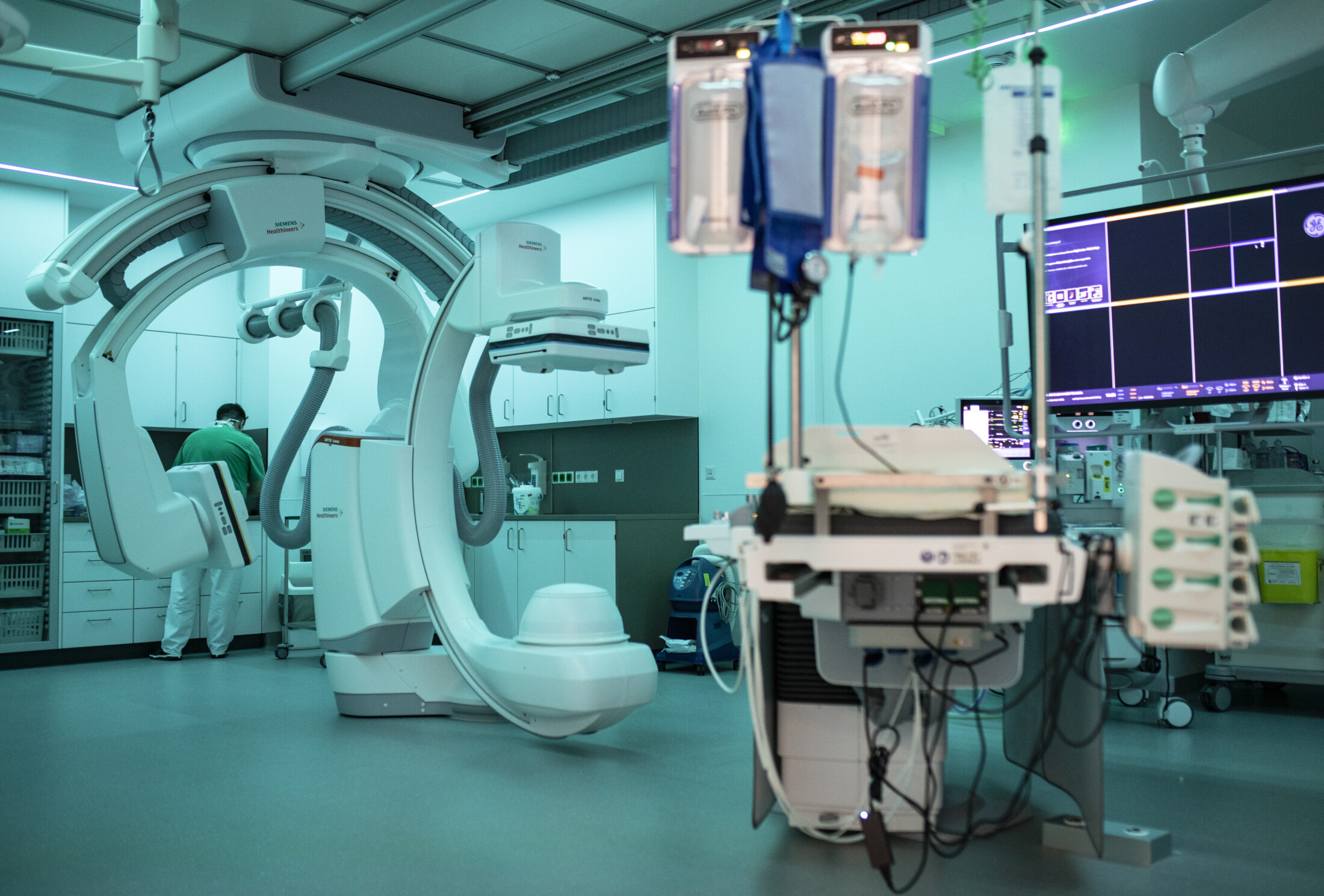 Medizinischer Raum mit einer Schrankwand im Hintergrund, davor ein sehr großes medizinisches Gerät. Rechts medizinische Instrumente zur Überwachung der Vitalfunktion. Vor der Schrankwand steht eine medizinische Fachkraft.