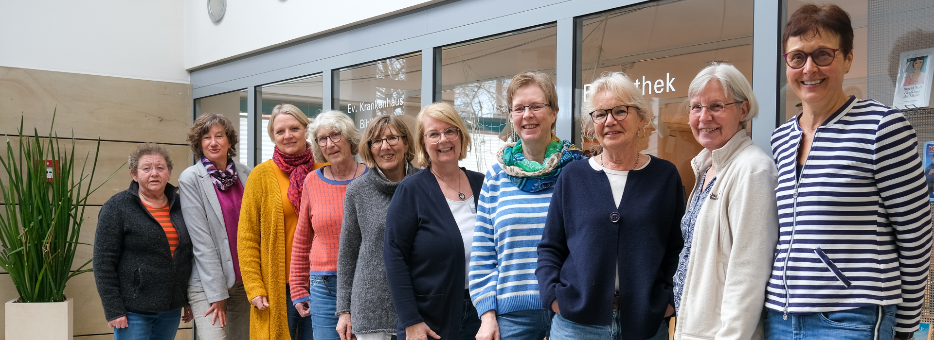 Gruppe von 10 weiblichen Personen in Alltagskleidung vor Glasscheiben mit der Aufschrift Bibliothek.