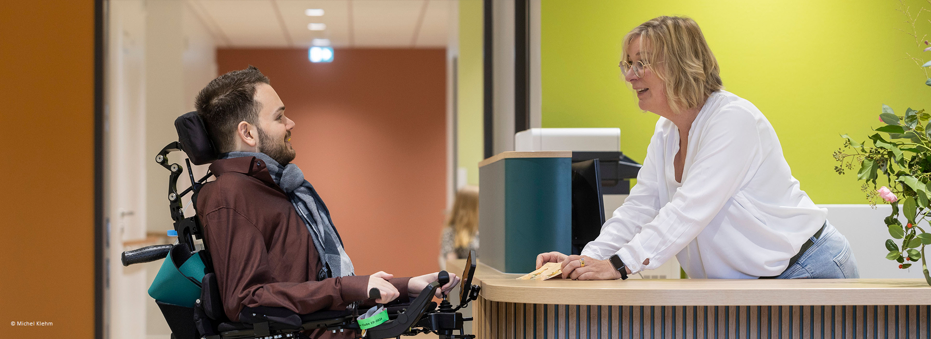 Mann in einem Rollstuhl und eine Frau hinter einem Tresen schauen sich lächelnd an.