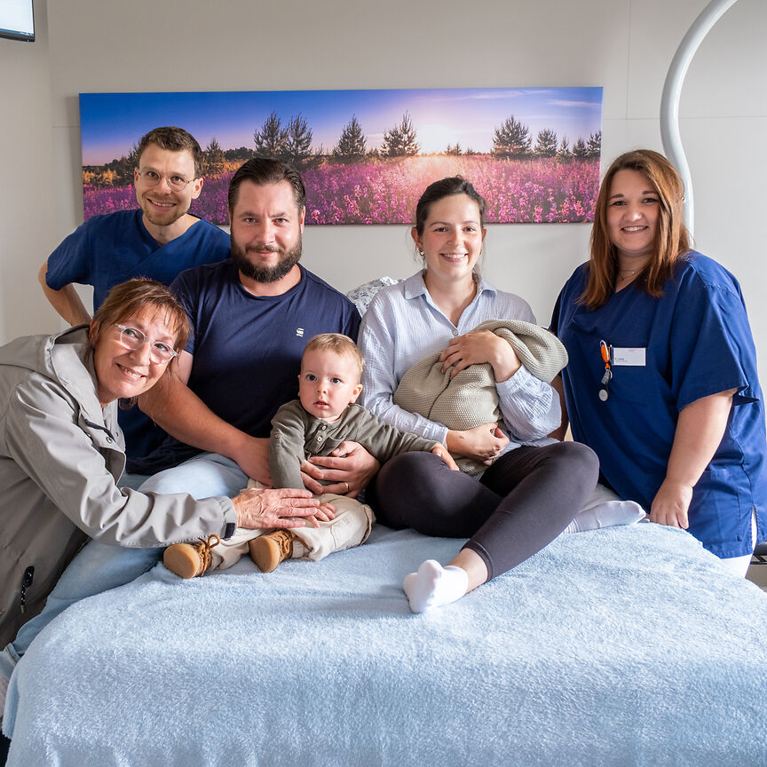 Fünf erwachsene Menschen von denen zwei einen blauen Kasack tragen. Ein Mann, eine Frau, die ein Säugling im Arm hält und ein kleiner Junge sitzen auf einem Bett. Links ist eine Frau, die zum Paar auf dem Bett gehört,