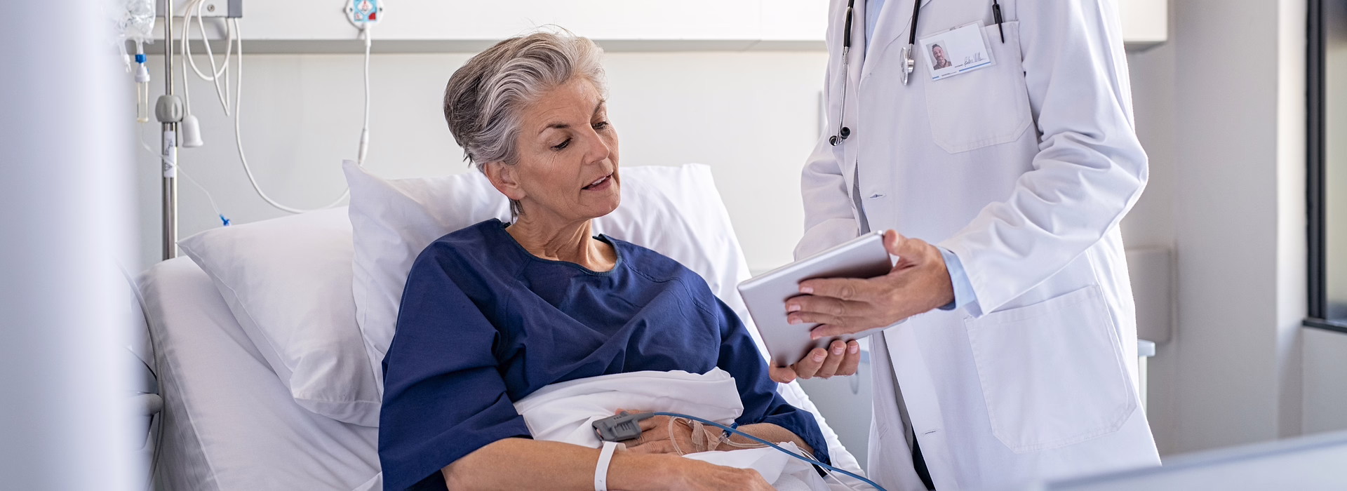 Frau in einem Bett. Eine Person in weißem Kittel hlt ihr ein Tablet hin.